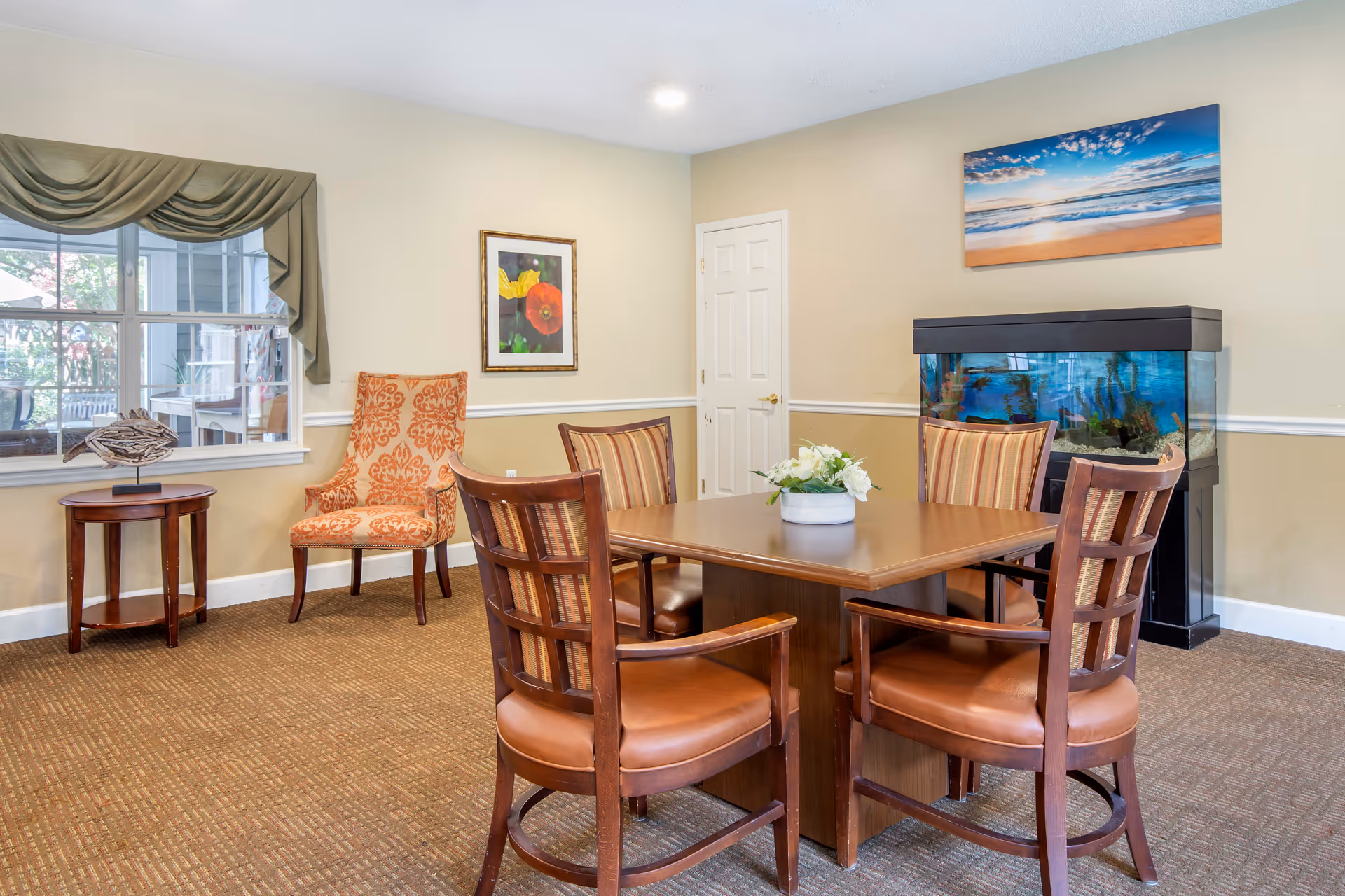 A cozy interior room with a square wooden table surrounded by four wooden chairs with cushioned seats and striped backs. There is an orange patterned armchair next to a small round wooden side table with a decorative wooden bird sculpture. A large window with green drapes lets in natural light. On the wall, there is a framed picture of flowers and a large aquarium beneath a beach sunset painting. The room has beige walls with white trim and a brown carpeted floor.