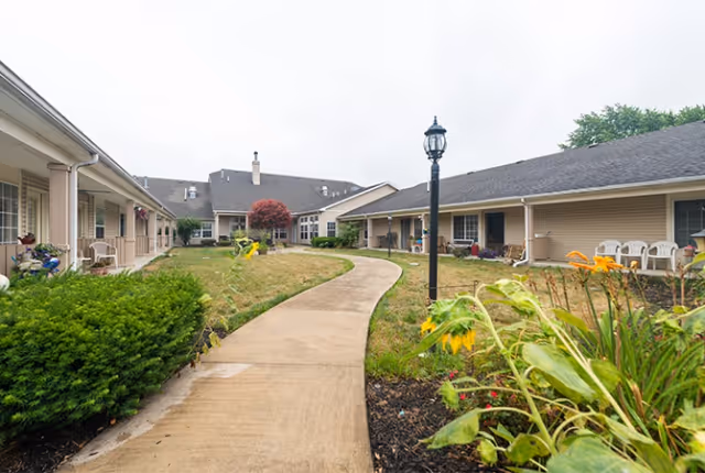 Curved concrete pathway through a landscaped courtyard between single-story senior living buildings with covered walkways and outdoor chairs.