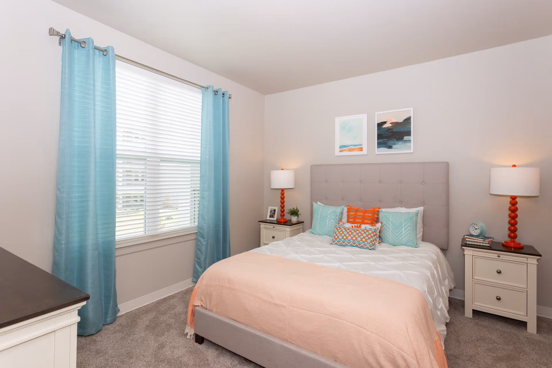A neatly arranged bedroom with a bed featuring a tufted gray headboard, white bedding, and decorative pillows in teal and orange. There are two white nightstands on either side of the bed, each with a lamp with a red base. A window with teal curtains and white blinds is on the left wall, and two abstract paintings hang above the bed.