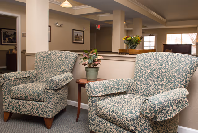 Two patterned upholstered armchairs flanking a small wooden side table with a potted plant in a quiet senior living common area.