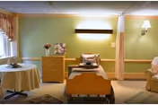 A senior living private room with an adjustable bed, dresser, armchair, privacy curtain, and pastel green walls.