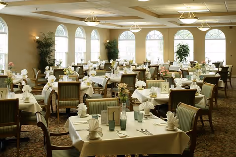 Spacious dining room with many neatly set tables, green-upholstered chairs, and arched windows letting in natural light.
