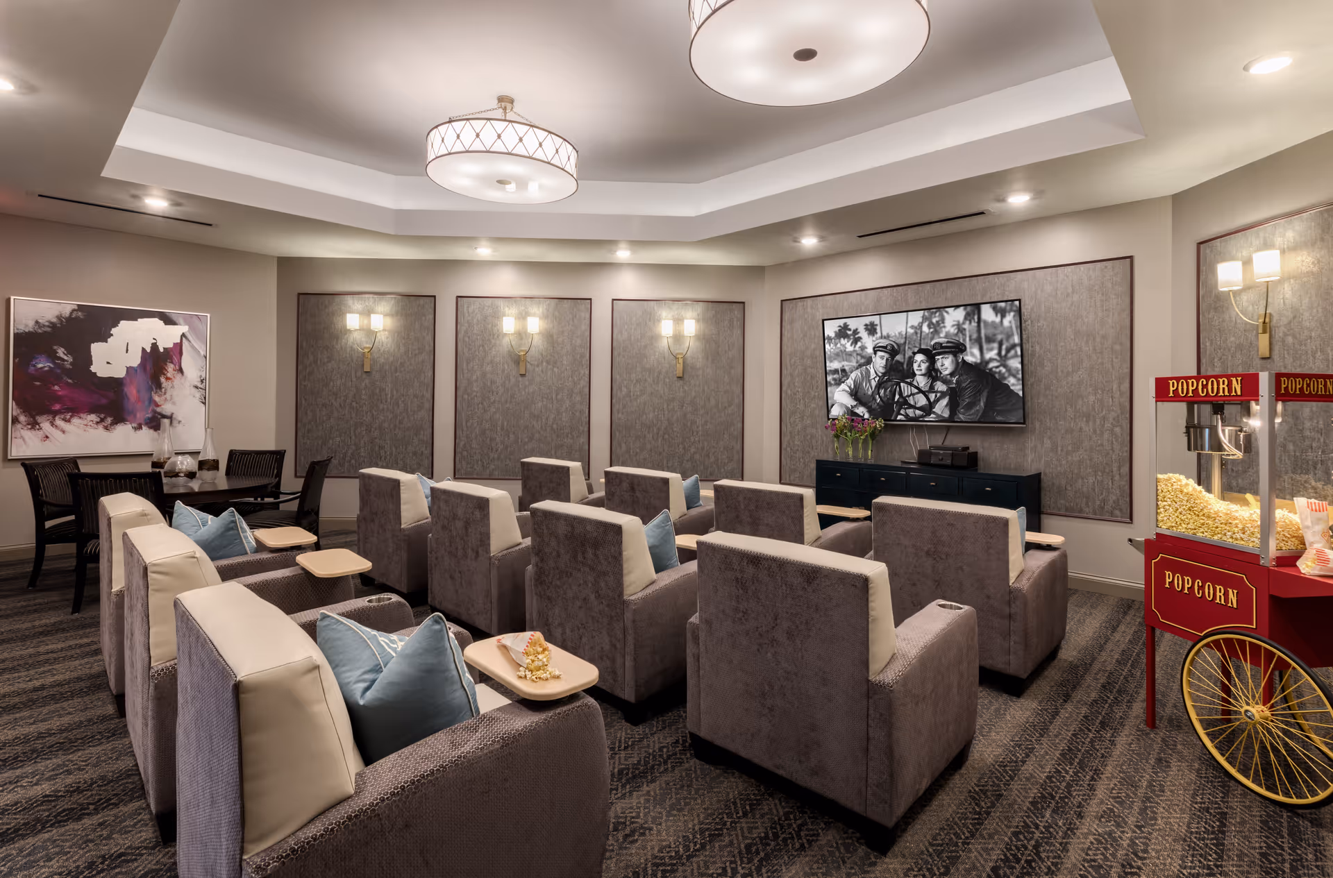 A cozy movie theater room with multiple rows of plush armchairs, each equipped with a small wooden tray. A large flat-screen TV is mounted on the wall showing a black and white movie scene. To the right, there is a vintage-style popcorn machine filled with popcorn. The room has soft lighting with wall sconces and ceiling fixtures, and a small table with chairs is visible in the back corner.