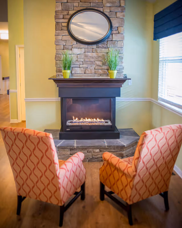 Two patterned armchairs facing a modern gas fireplace with a stone surround and a wooden mantel. Above the mantel is a round mirror, and two small green plants in yellow pots are placed on the mantel. The room has light yellow walls and a window with blinds on the right side.
