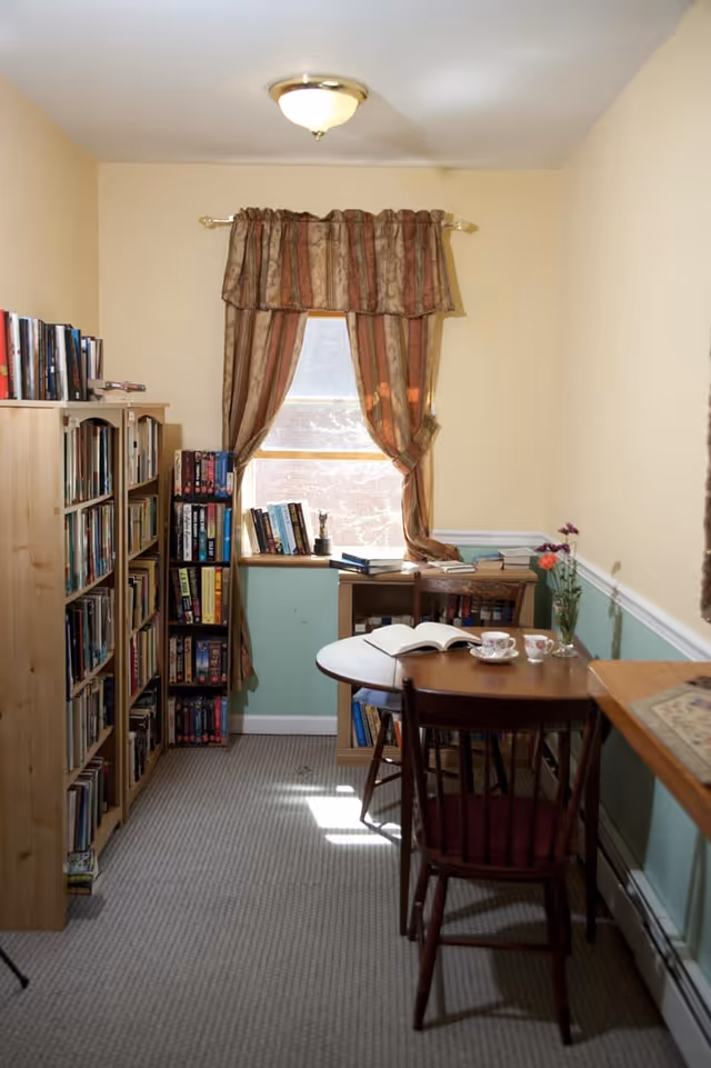 A cozy reading room with bookshelves filled with books along the left wall, a window with patterned curtains letting in natural light, a small wooden table with two chairs, an open book, two teacups, and a vase with flowers on the table. The walls are painted beige and light green with a white chair rail.