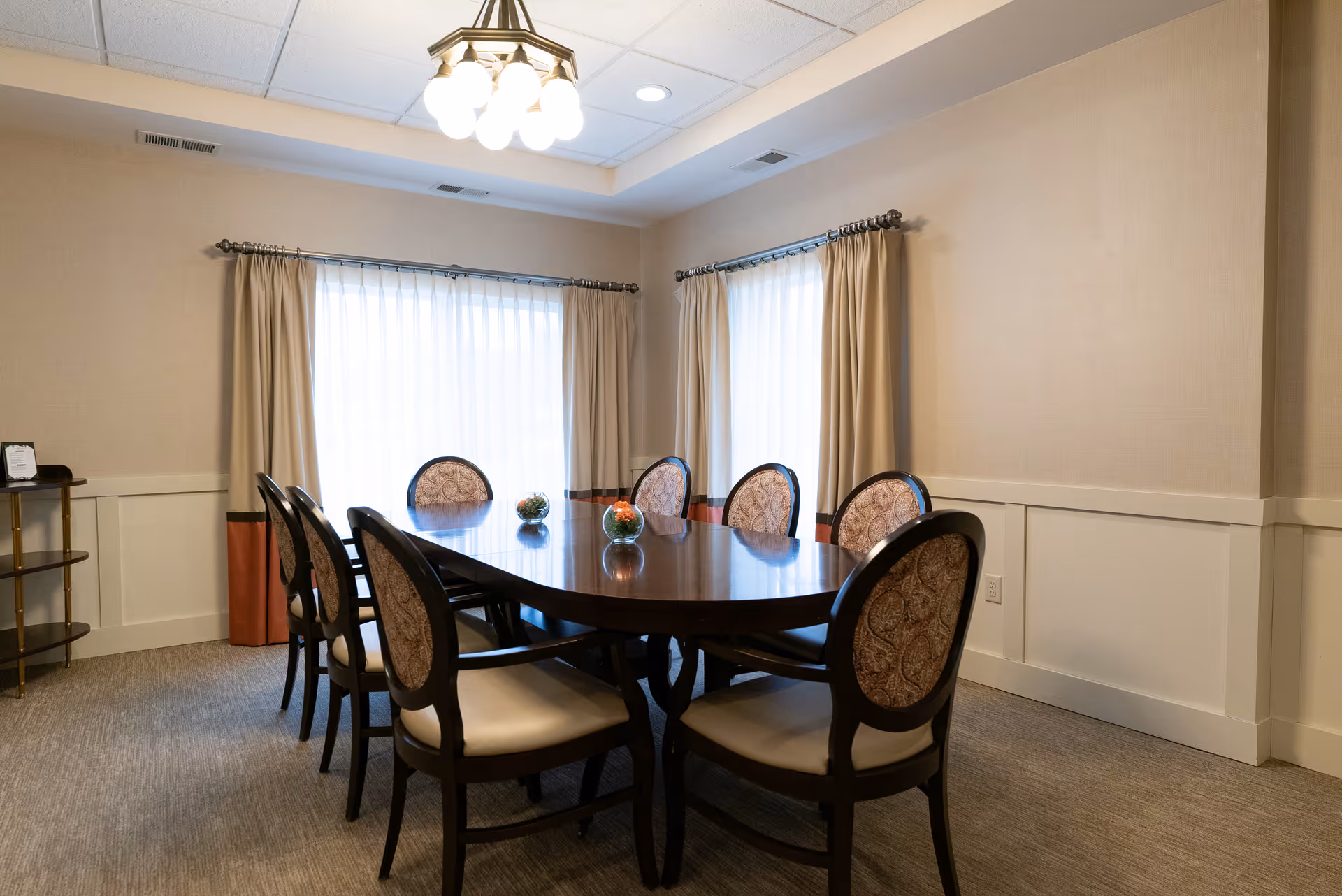 A dining room with a polished wooden table surrounded by eight upholstered chairs. The room has beige walls with white wainscoting, two windows with beige curtains, and a ceiling light fixture with multiple bulbs. There are three small decorative glass bowls with flowers on the table.