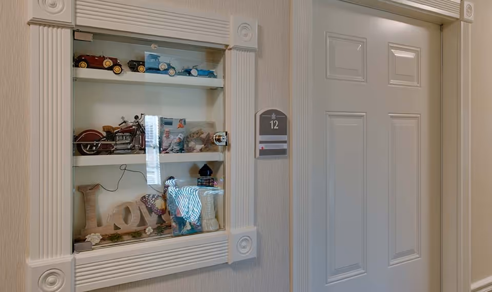 A white door labeled with the number 12 next to a built-in glass display case containing miniature model cars, a motorcycle, a photo, decorative letters spelling 'Love', and other small decorative items.