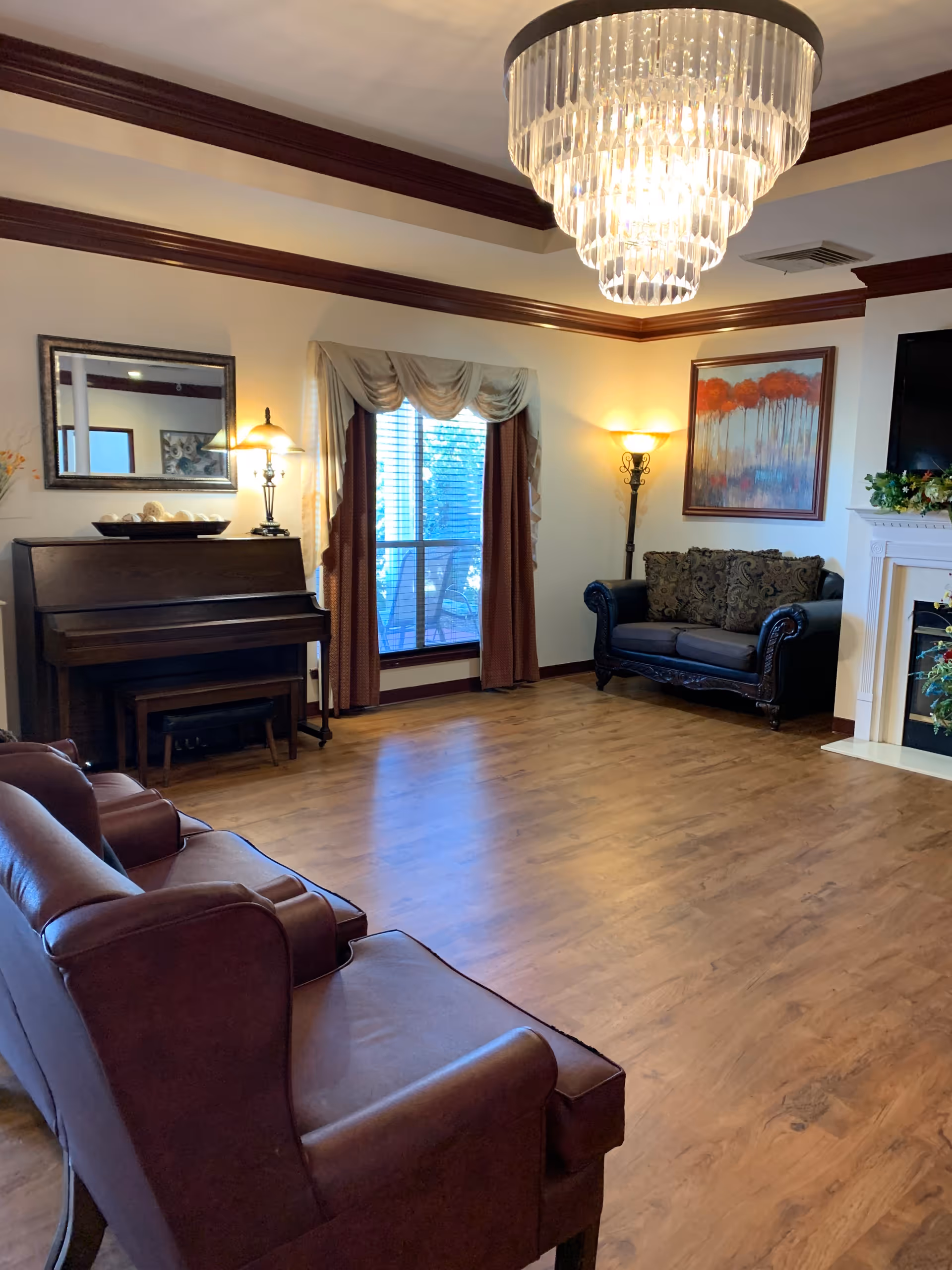 A cozy living room with wooden flooring, featuring two brown leather armchairs in the foreground, a dark wood piano against the left wall with a mirror and lamp above it, a window with draped curtains in the center, and a dark upholstered loveseat with patterned cushions near a white fireplace on the right. A large crystal chandelier hangs from the ceiling, and a colorful painting is mounted on the wall above the loveseat.
