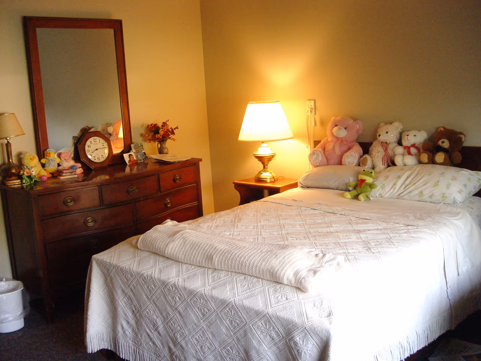 A cozy bedroom with a neatly made bed covered in a white textured blanket and several stuffed animals lined up on the headboard. A wooden nightstand with a lit lamp is beside the bed. Across from the bed is a wooden dresser with a large mirror, a clock, a small lamp, and various decorative items including stuffed animals and a photo frame.