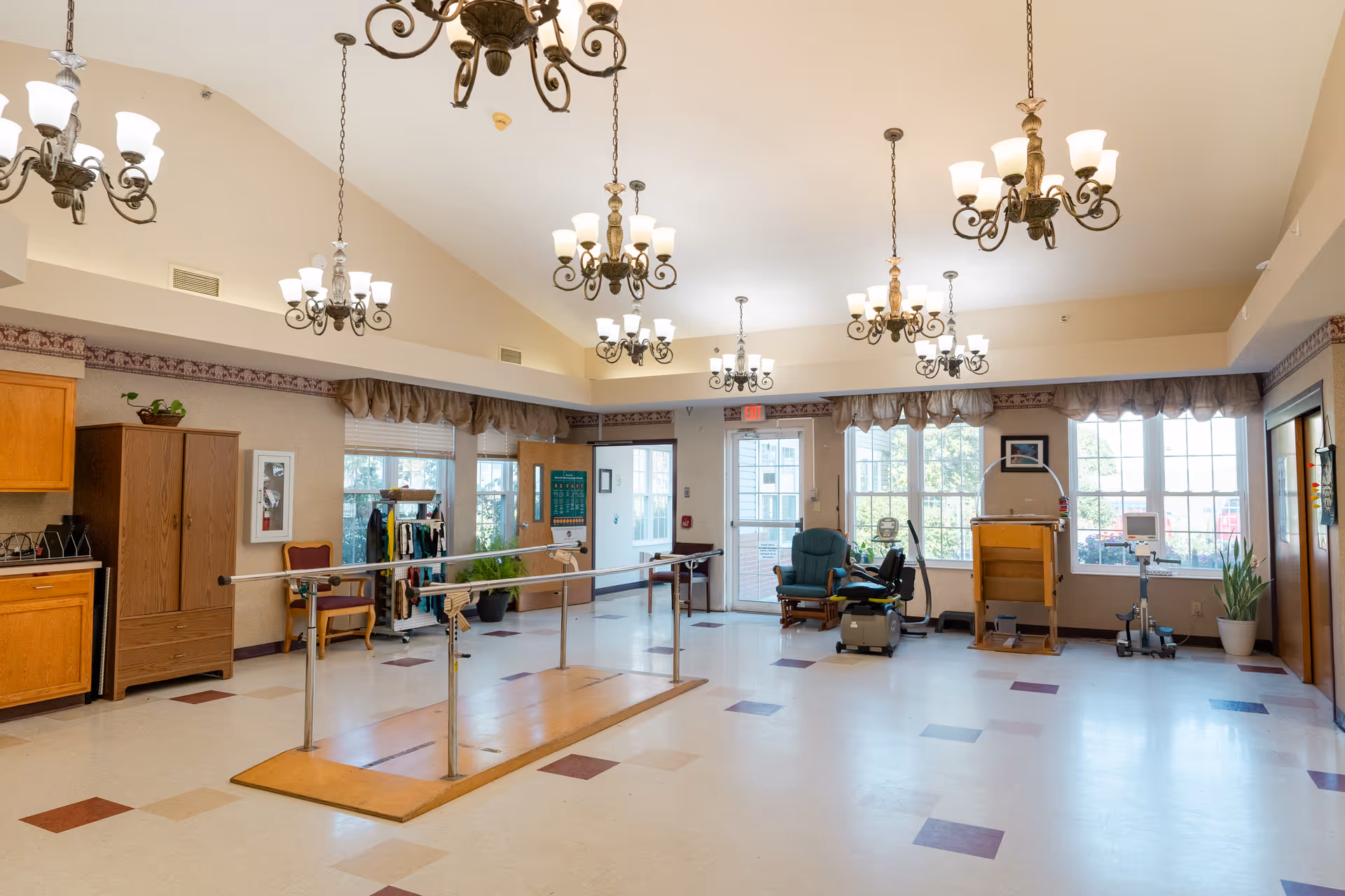 A spacious rehabilitation room with parallel bars for walking exercises in the center, several chandeliers hanging from the ceiling, large windows letting in natural light, exercise equipment including a stationary bike and a chair, wooden cabinets, and plants placed around the room.