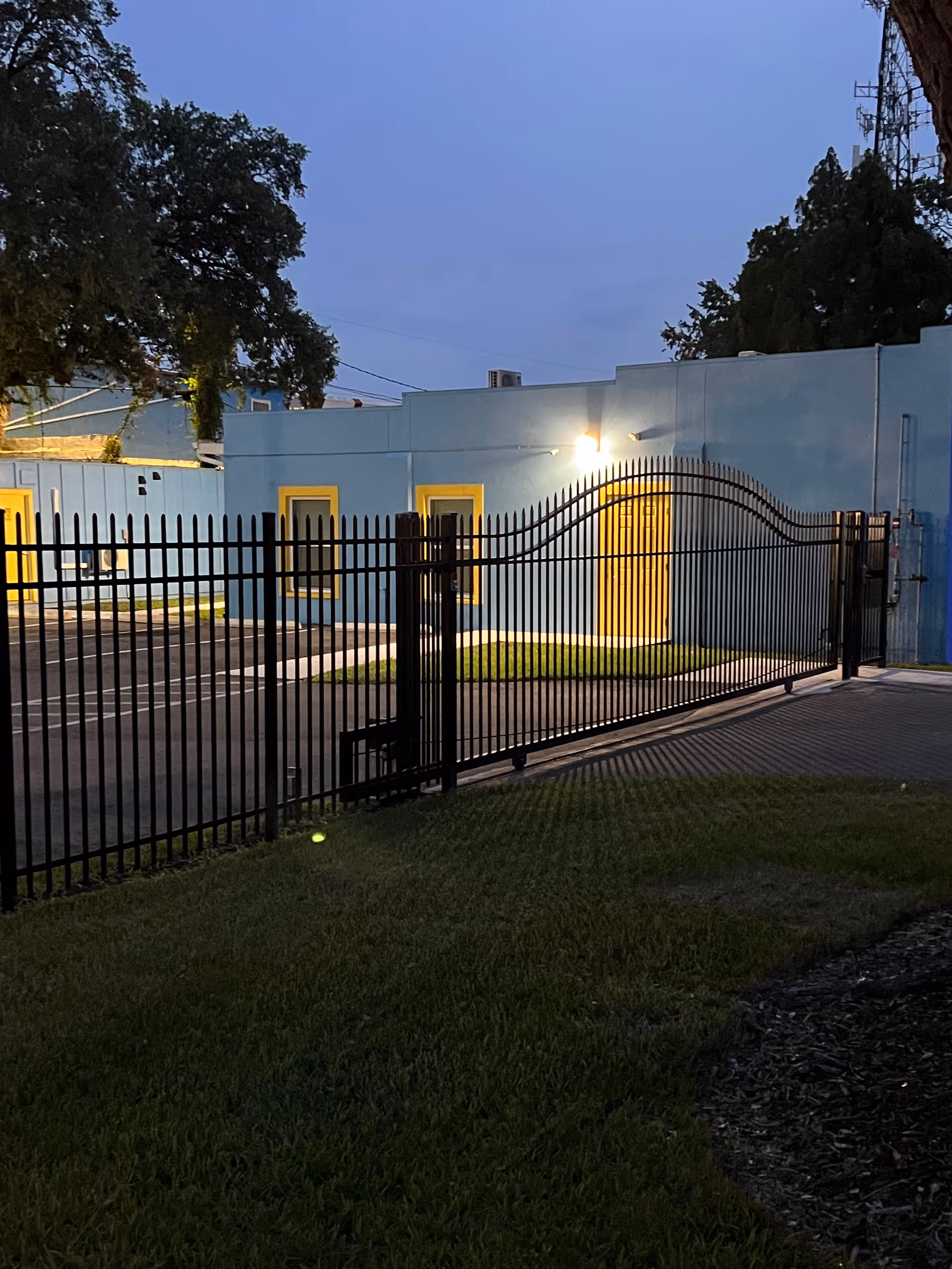 Gated front of a single-story light-blue building with yellow-trimmed doors and windows illuminated at dusk