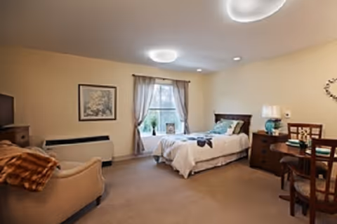 A cozy bedroom in an assisted living facility featuring a single bed with white bedding and a blue throw pillow, a wooden nightstand with a lamp, a small round table with two chairs, a beige armchair with a brown throw blanket, a window with curtains, and a framed picture on the wall.