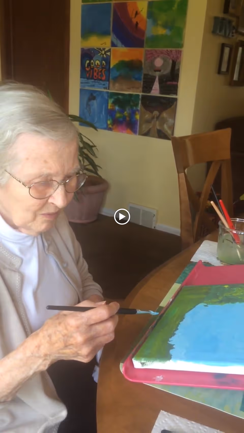 An elderly woman with glasses is sitting at a table painting on a canvas. Behind her on the wall are colorful paintings arranged in a grid. The room has warm lighting and wooden furniture.