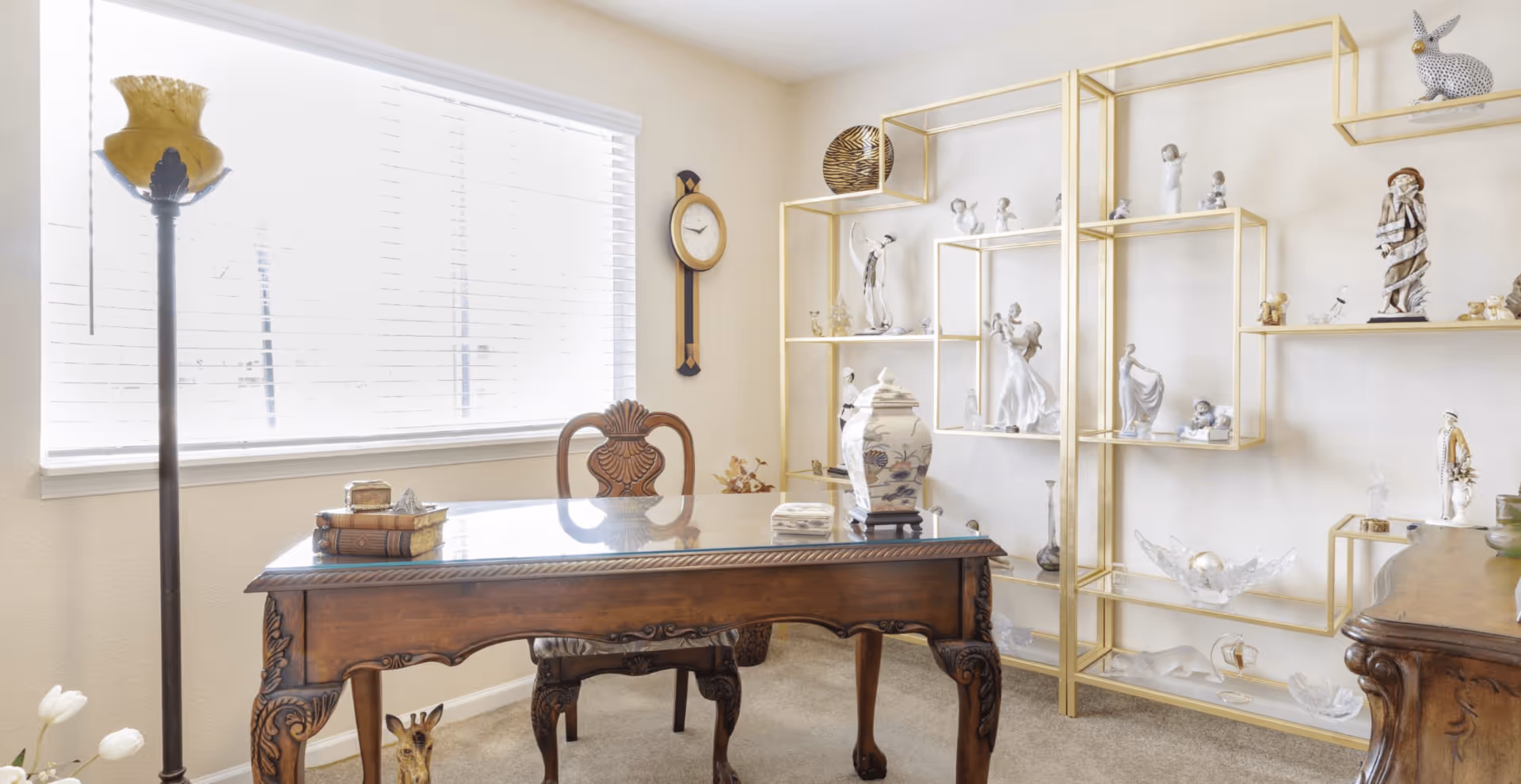 Sunlit interior room with an ornate wooden desk, gold-framed display shelves filled with figurines, a floor lamp, and a wall clock.