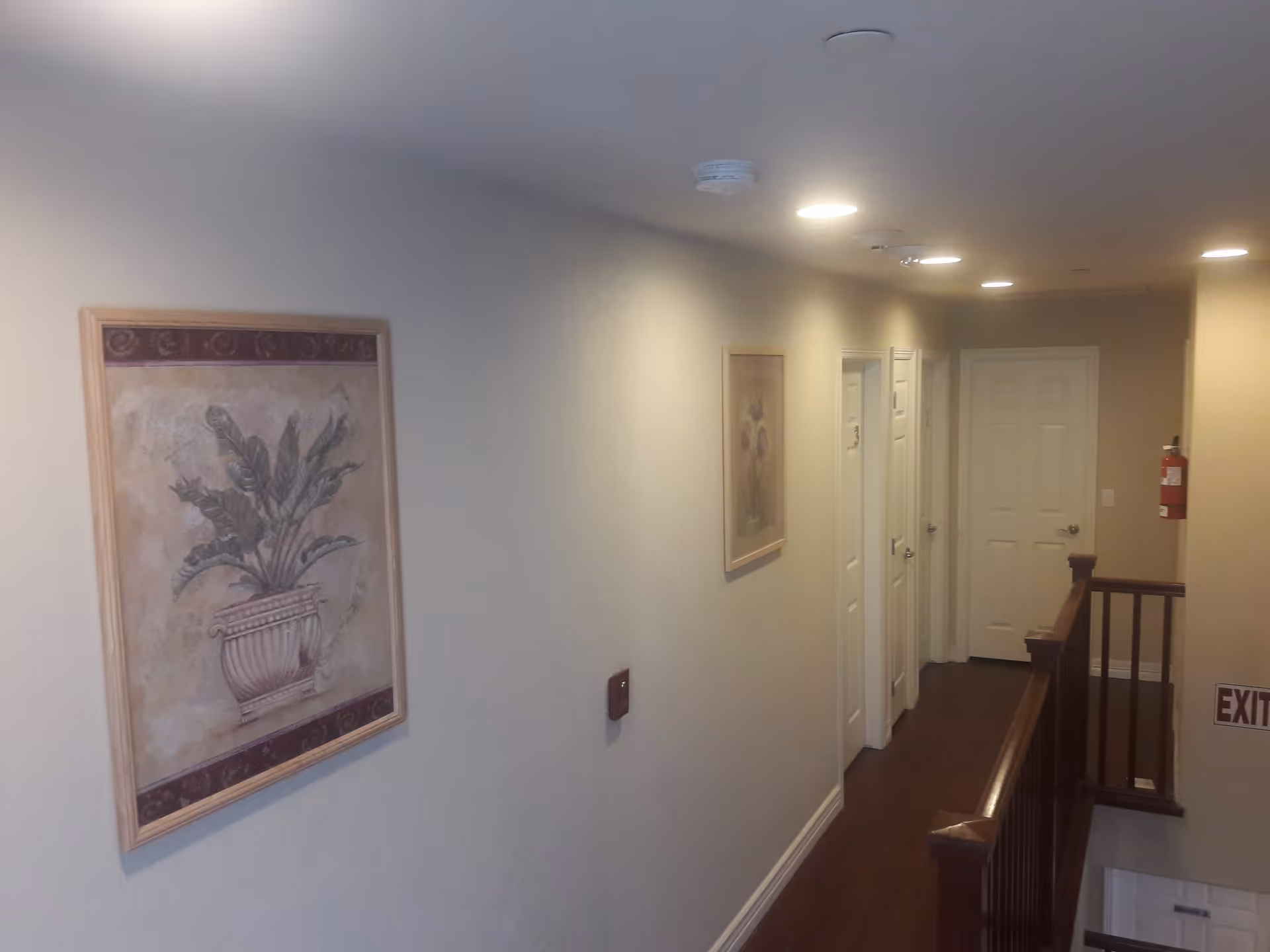 Interior hallway of Auburn View Assisted Living with framed artwork on the walls, multiple closed white doors, wooden handrails along the staircase, and ceiling lights illuminating the space.
