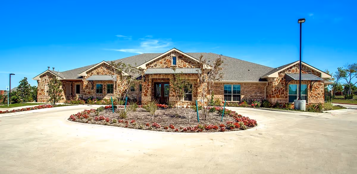Front exterior view of a single-story stone building with a circular driveway and landscaped garden area in front, under a clear blue sky.