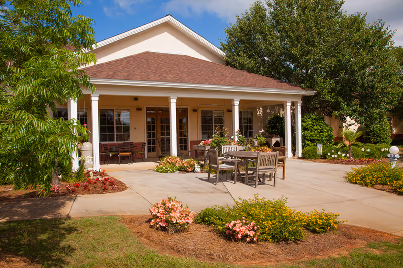 Front entrance of a residential building with a covered porch, outdoor tables and chairs, and landscaped flower beds.