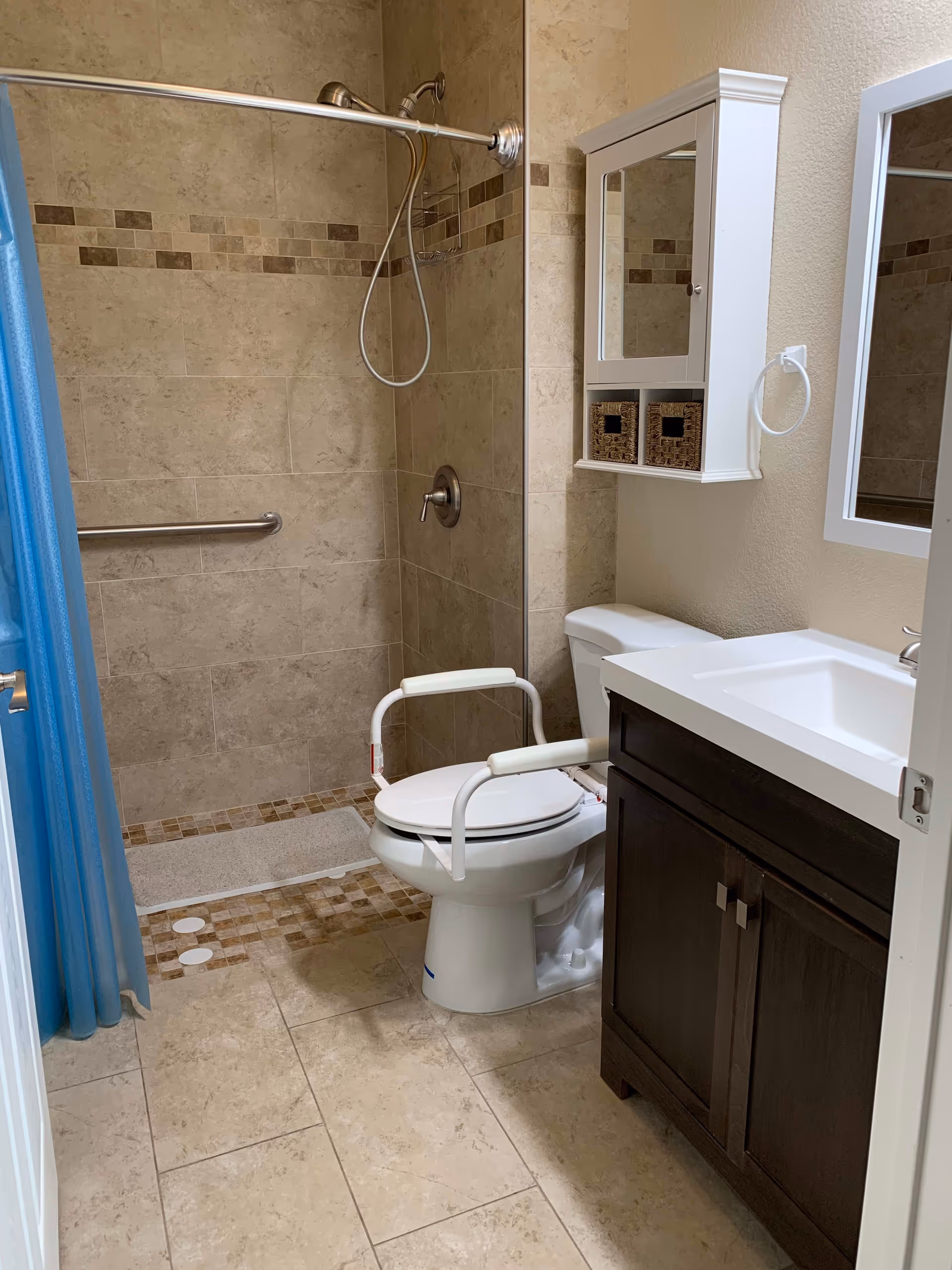 A senior-friendly bathroom featuring a walk-in tiled shower with a handheld showerhead and grab bar, a toilet with white safety rails, a dark wood vanity with a white sink, a wall-mounted cabinet with two wicker baskets, and a mirror above the sink. The floor is tiled in beige tones, and a blue shower curtain is partially visible.