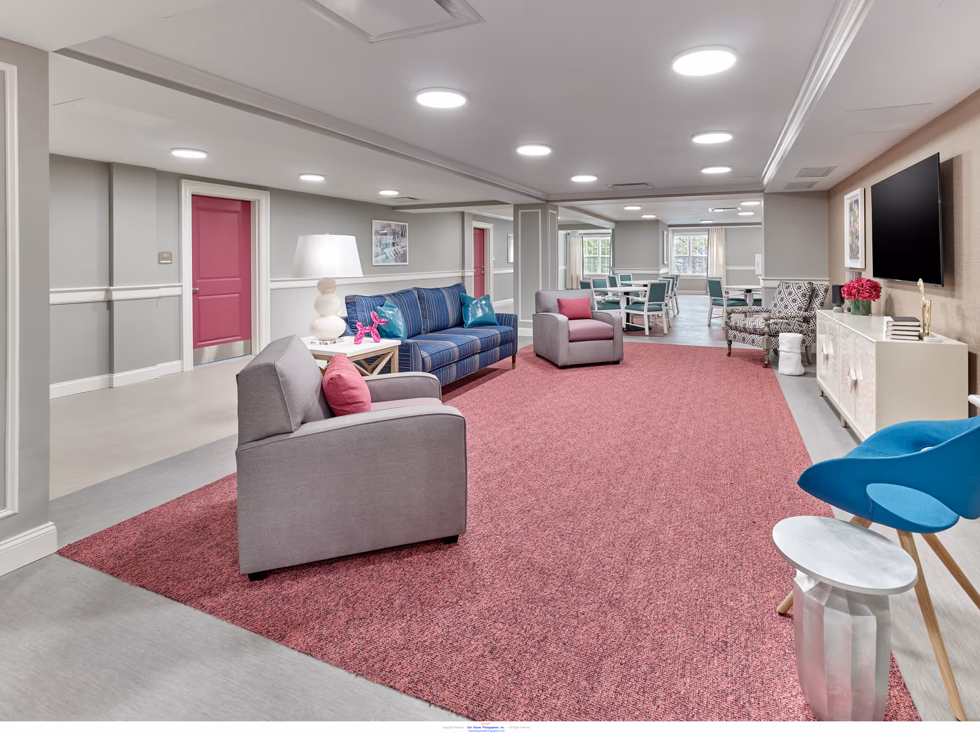 Bright communal lounge with sofas, armchairs, a TV, and a dining area in the background.