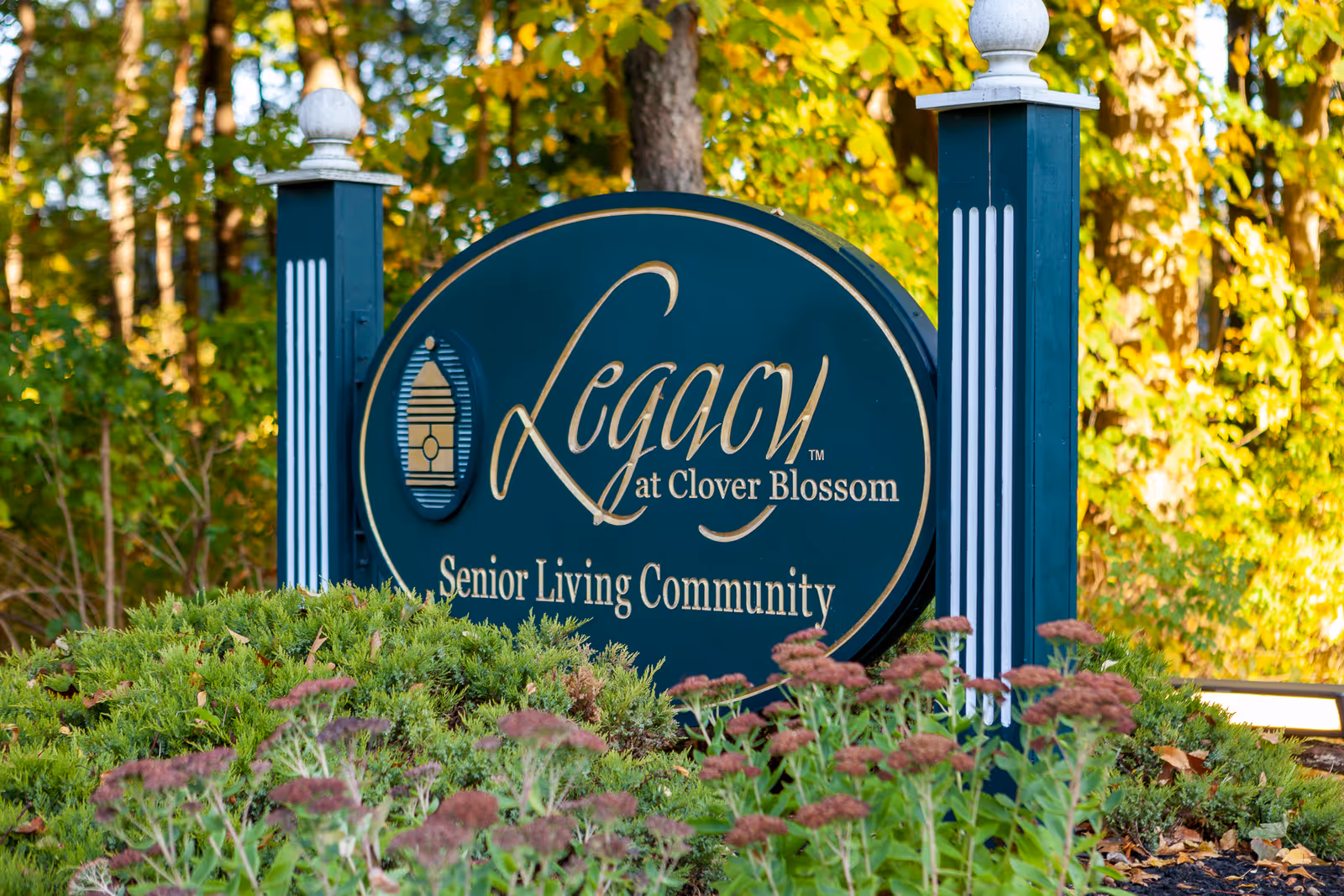 A large oval sign for Legacy at Clover Blossom Senior Living Community, mounted between two decorative posts with white finials, surrounded by greenery and flowers with trees in the background.