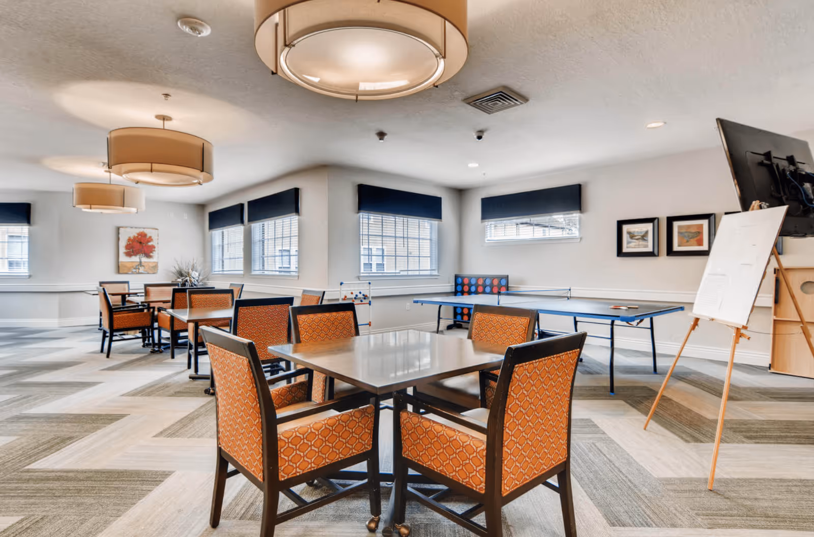 A bright activity room with multiple tables and orange patterned chairs, large windows with dark valances, ceiling lights, a ping pong table, a large TV on a stand, and an easel with a whiteboard.