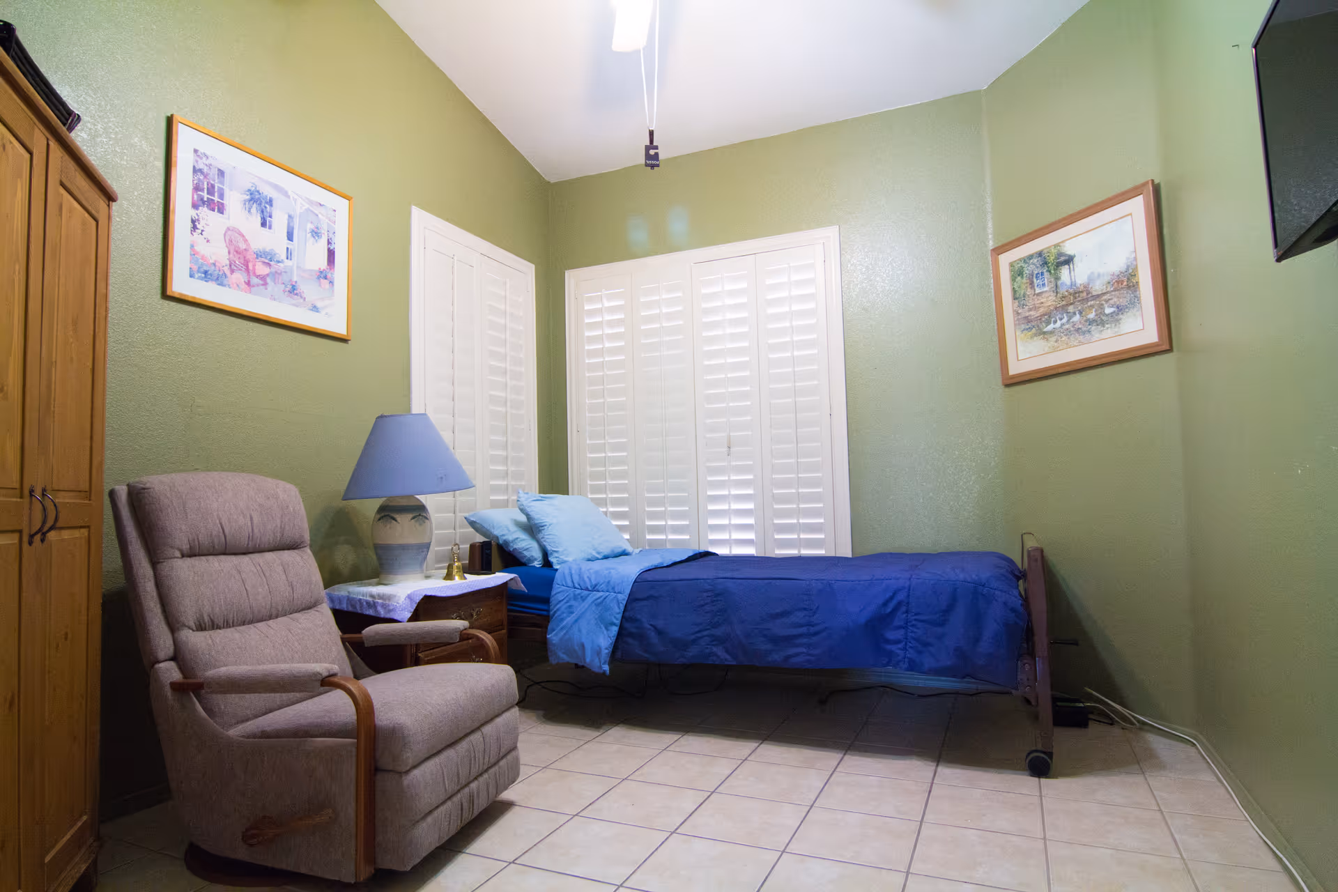 A small bedroom with green walls and tiled floor featuring a single bed with blue bedding, a beige cushioned armchair, a wooden nightstand with a blue lamp, and two framed pictures on the walls. White plantation shutters cover the windows, and a flat-screen TV is mounted on the wall.