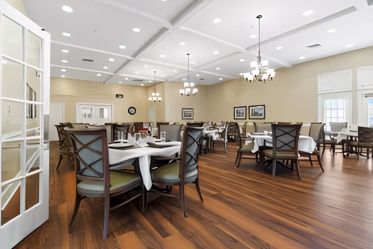 A spacious dining room in Carillon Assisted Living of Hendersonville with multiple tables covered in white tablecloths and set with plates and glasses. The room features wooden flooring, beige walls, several chandeliers hanging from a white ceiling with recessed lighting, and large windows allowing natural light to enter.