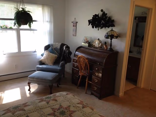 A cozy bedroom corner with a blue upholstered armchair and matching ottoman near a window with sheer white curtains. Next to the chair is a wooden roll-top desk with a wooden chair, decorated with a table lamp, clock, and small decorative items. A doorway to a bathroom is visible on the right side of the image.