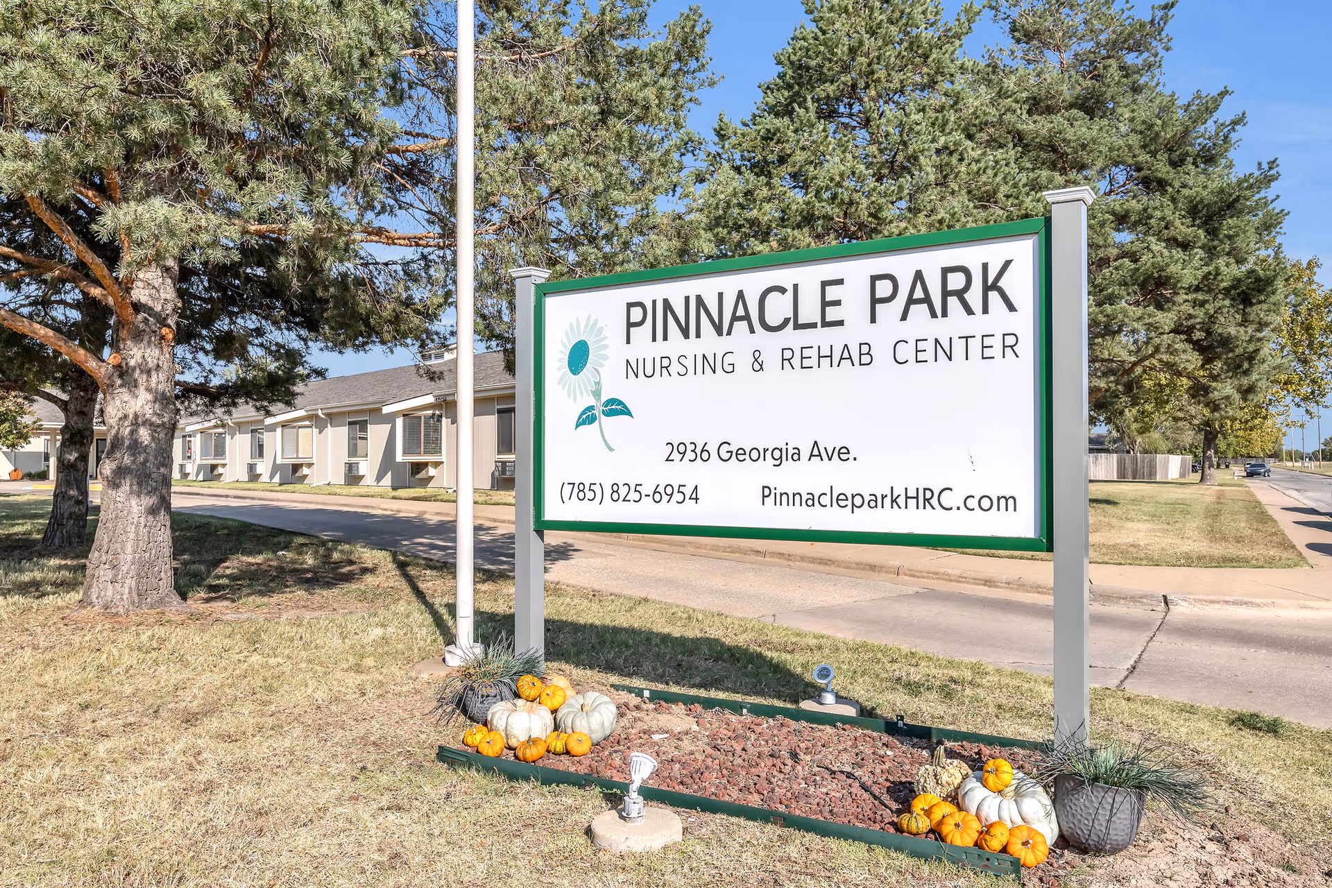 Outdoor view of Pinnacle Park Nursing & Rehab Center sign with address 2936 Georgia Ave and contact information. The sign is surrounded by small pumpkins and landscaping rocks, with trees and part of the building visible in the background.