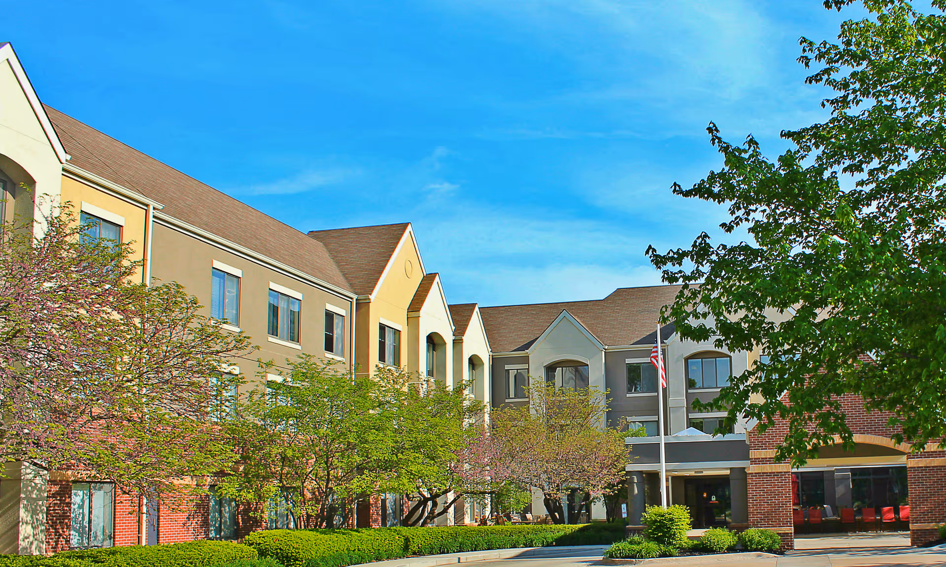 Exterior view of a multi-story retirement facility building with a mix of beige, gray, and brick walls, surrounded by green trees and bushes under a clear blue sky. An American flag is visible near the entrance.
