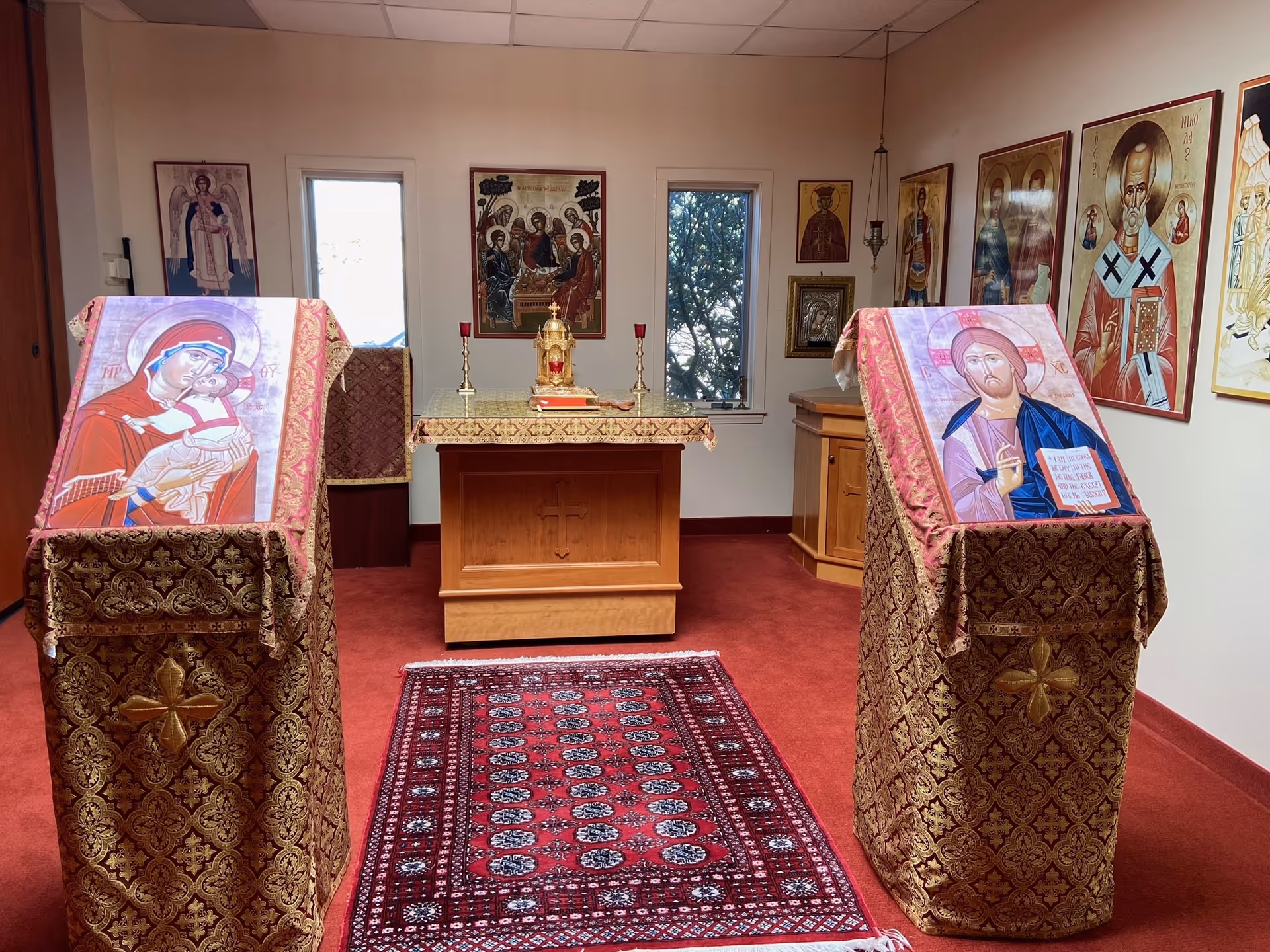 Interior of a small chapel room with religious icons and paintings on the walls. Two stands draped in ornate red and gold cloth hold large religious images, one of the Virgin Mary holding baby Jesus and the other of Jesus Christ. A wooden altar with a cross carved on the front is covered with a matching cloth and holds religious items including candles and a gold tabernacle. A red patterned rug lies on the floor in front of the altar.