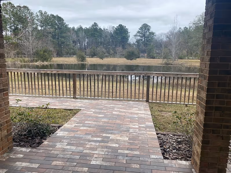 View from a covered brick patio looking out over a wooden railing towards a small pond and a grassy area with trees in the background under a cloudy sky.