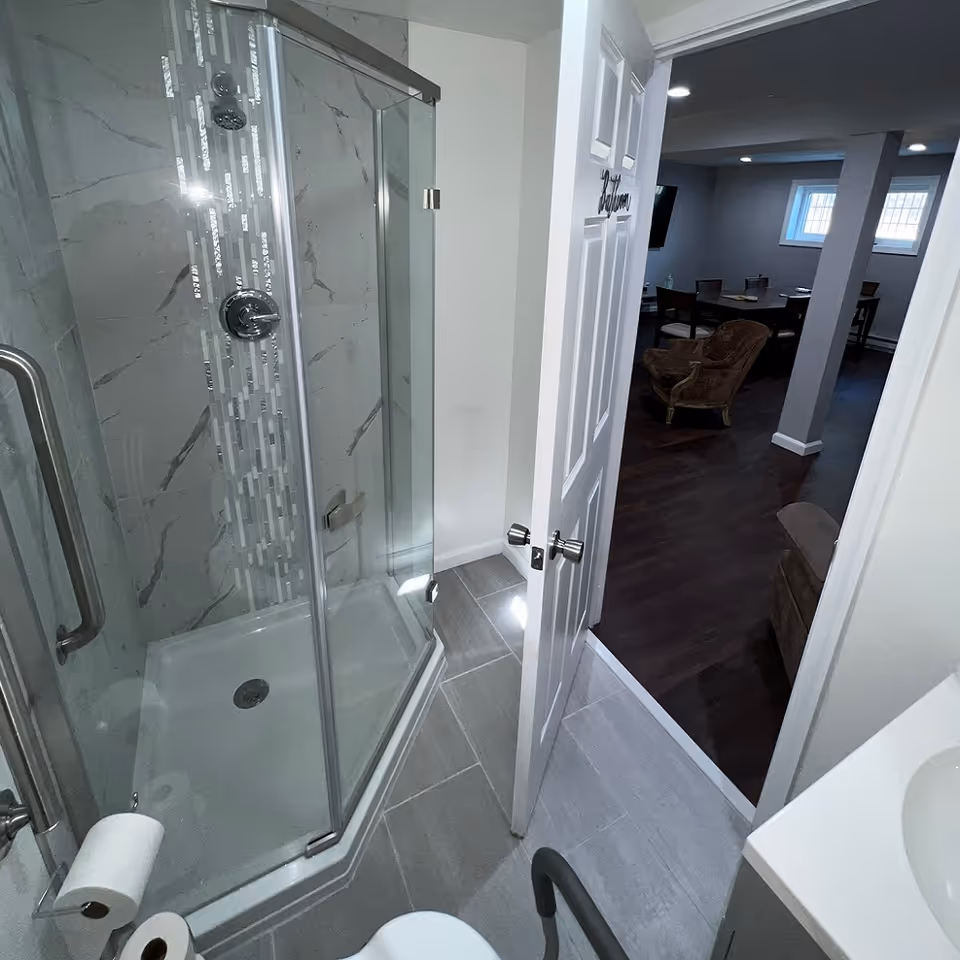 A modern bathroom with a glass-enclosed corner shower featuring marble-patterned tiles and a vertical strip of decorative tiles. The bathroom has gray floor tiles, a white sink countertop, and a toilet with a grab bar. The door is open, showing a glimpse of a living area with dark wood flooring, chairs, a table, and windows.