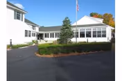 Front entrance of a white nursing home building with a circular driveway, landscaped shrubs, and an American flag.