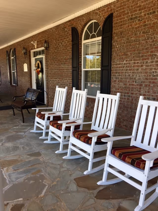 A covered porch area with a stone floor featuring four white wooden rocking chairs with striped cushions lined up in front of a brick wall with black shuttered windows and a black door decorated with a wreath. There is also a dark wooden bench near the door.