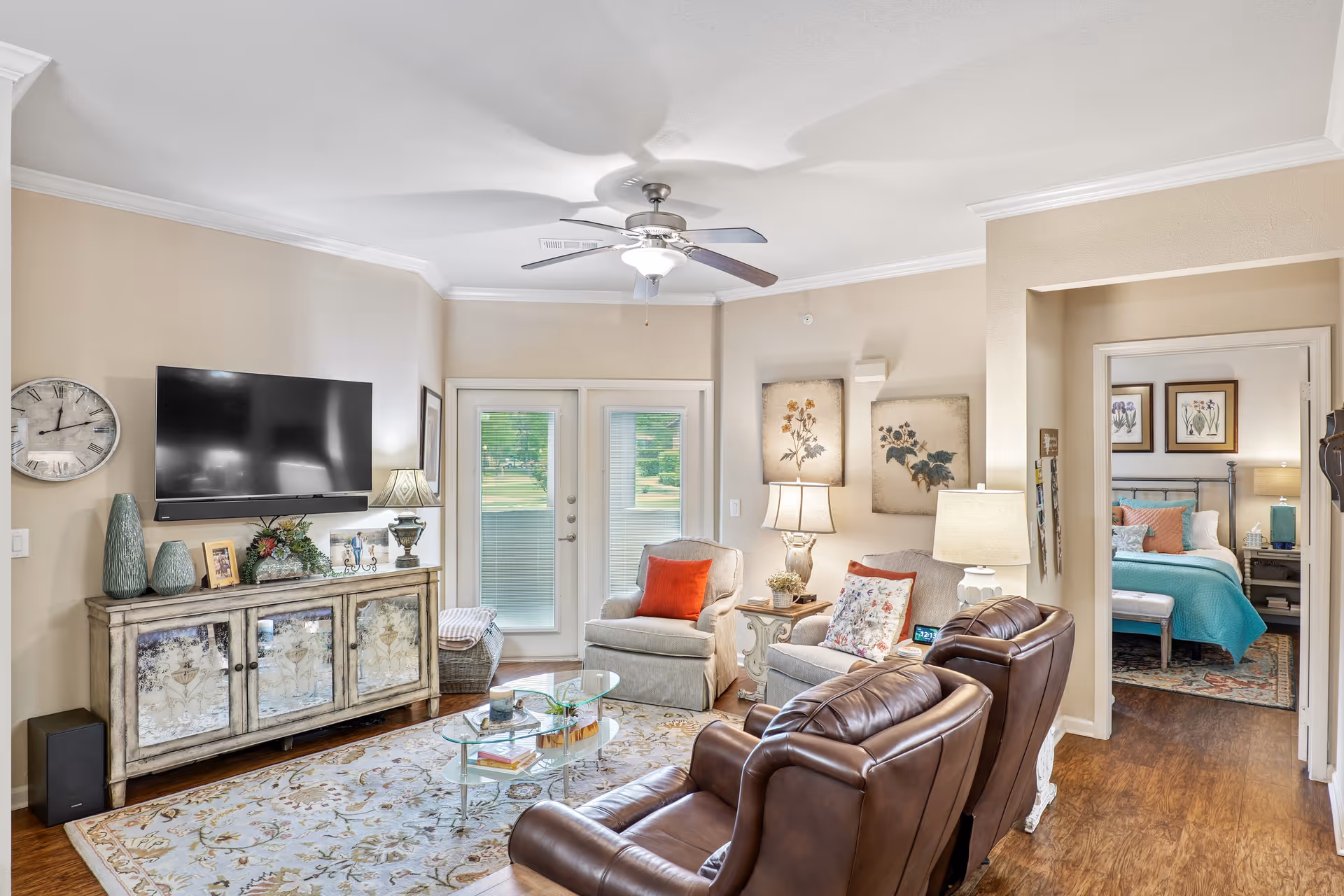 Bright furnished living room with leather recliners, armchairs, a wall-mounted TV on a console, glass coffee table, and a doorway to a bedroom.