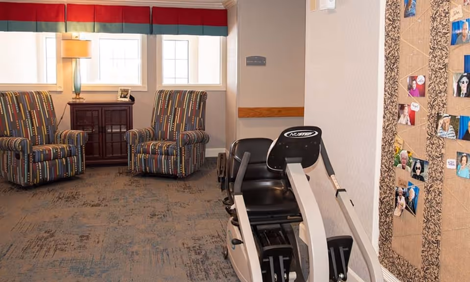 A cozy room with two patterned armchairs, a wooden cabinet with a lamp and picture frame, a recumbent exercise bike, and a bulletin board displaying various photos on the wall.