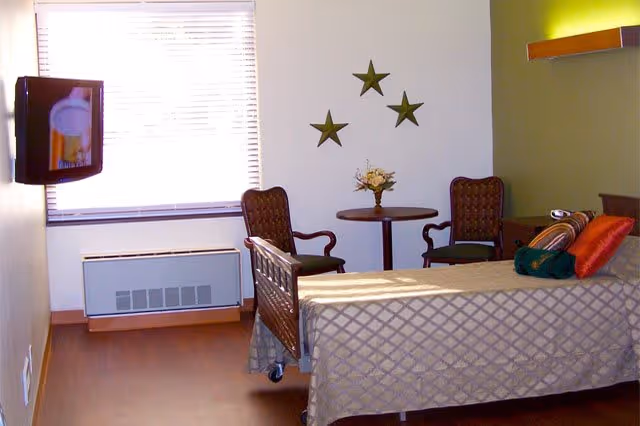 A well-lit bedroom in a healthcare facility featuring a single bed with a patterned bedspread and colorful pillows. There are two upholstered chairs and a small round table with a flower arrangement between them. Three decorative stars are mounted on the wall above the table. A wall-mounted television is positioned near a window with blinds, and a heating unit is located beneath the window.