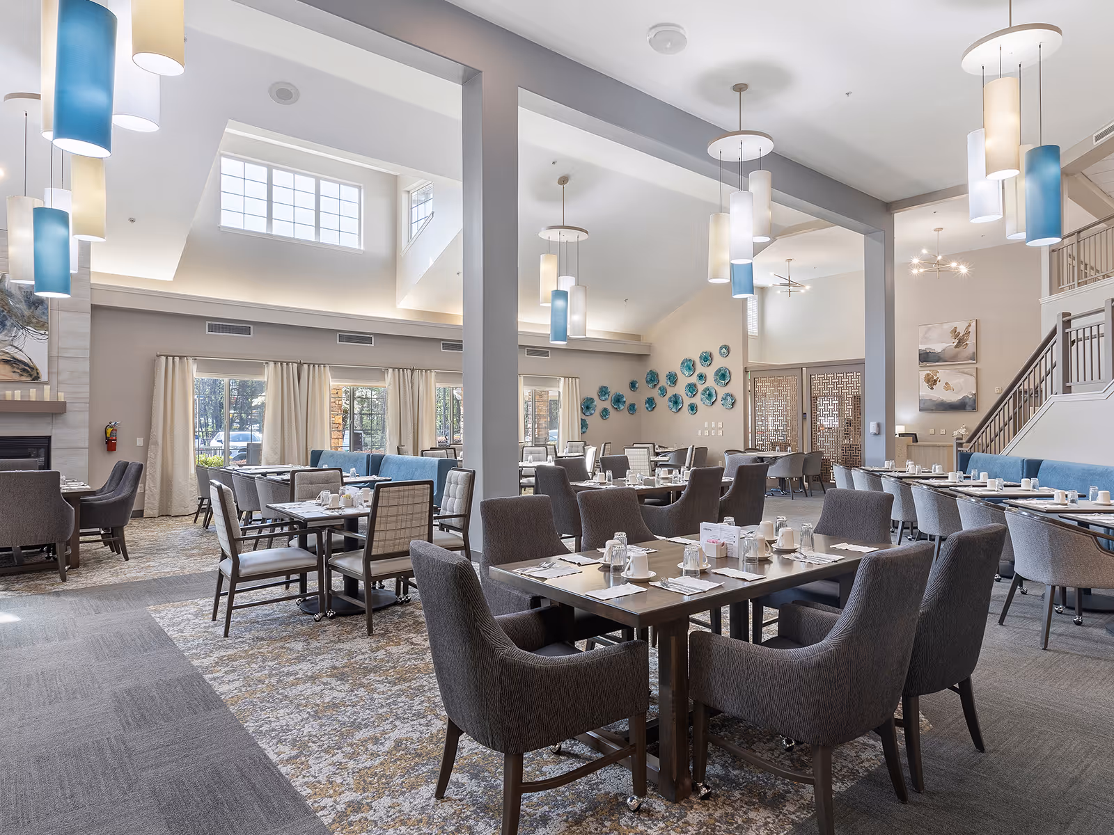 Spacious, well-lit dining room with multiple set tables and upholstered chairs under high ceilings and pendant lighting.