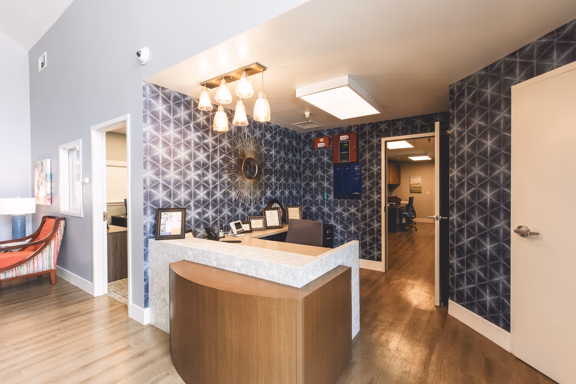 Reception area with a modern desk and chair, geometric patterned dark blue wallpaper, a decorative sunburst mirror, and pendant lights hanging from the ceiling. There are two open doorways leading to office spaces, a wooden floor, and a red upholstered chair with a lamp on the left side.