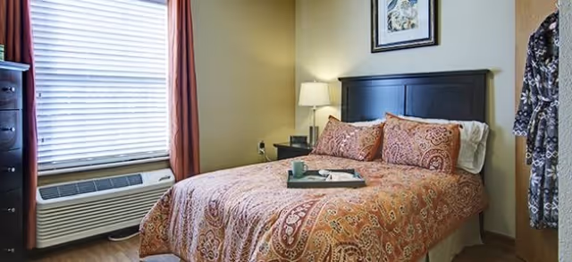 A cozy bedroom with a double bed covered in a patterned orange and red bedspread, two matching pillows, a tray with a cup and a book on the bed, a nightstand with a lamp, a window with blinds and red curtains, a dresser, and a robe hanging on the wall.