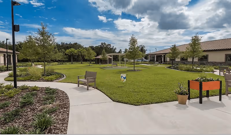 A well-maintained outdoor garden area with green grass, young trees, and a paved walkway. There are benches and planters with flowers along the path, and buildings with tiled roofs surround the garden. The sky is partly cloudy.