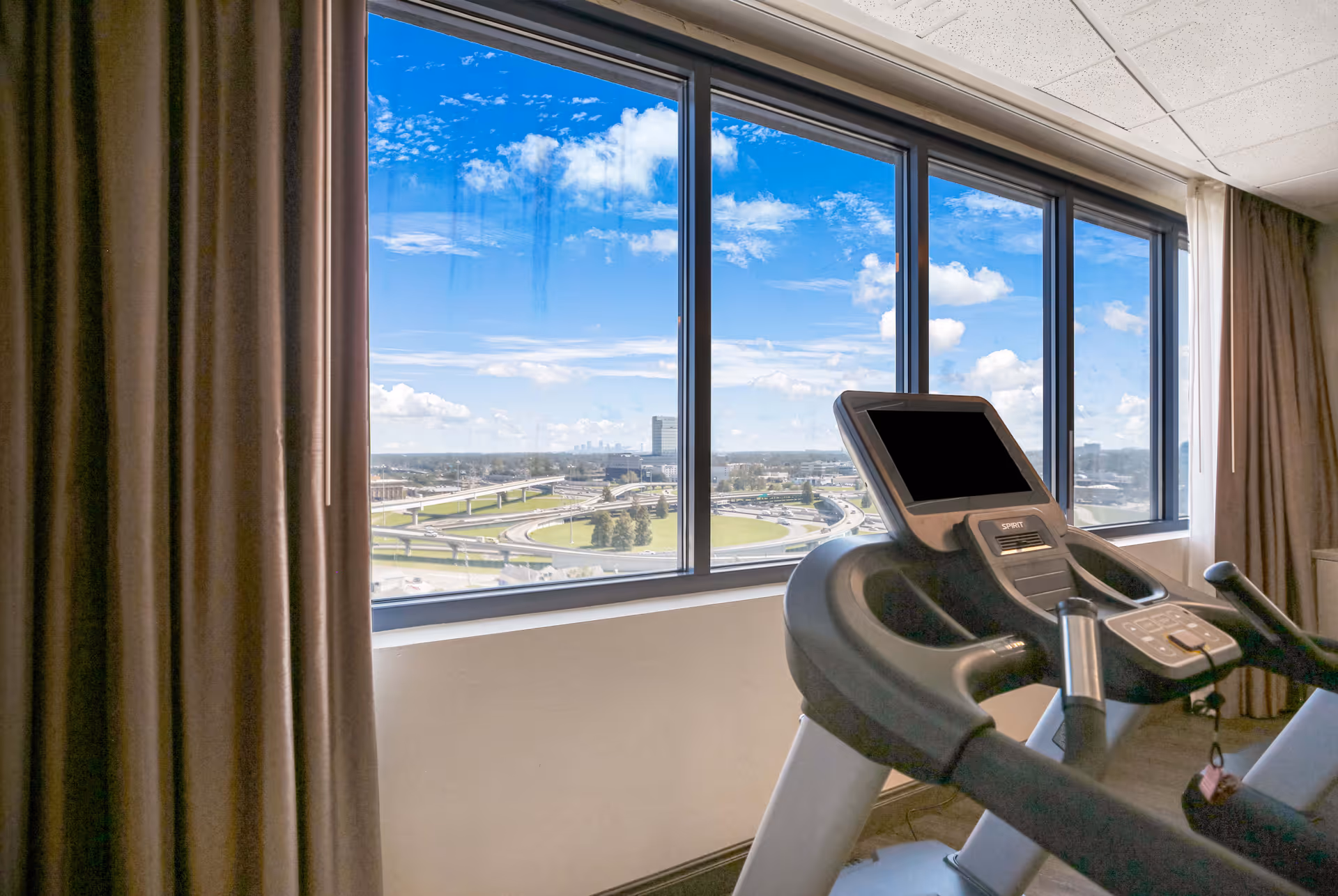 Indoor exercise room with a treadmill positioned near large windows offering a view of a highway interchange and a cityscape under a blue sky with scattered clouds. Brown curtains frame the windows.