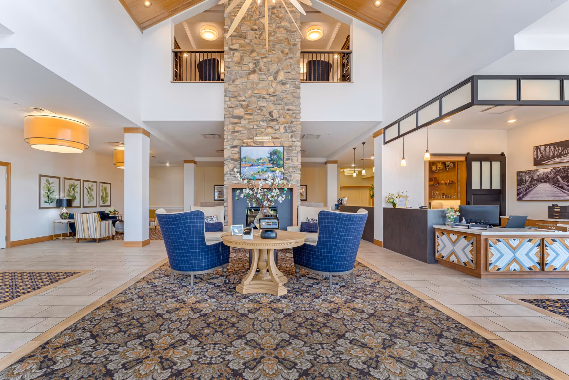 A spacious and well-lit senior living facility lobby with a high ceiling and a large stone fireplace in the center. In front of the fireplace, there is a round wooden table with a flower arrangement and two blue upholstered chairs. To the right, there is a reception desk with a computer and decorative panels. The floor is tiled with a large patterned rug in the seating area. The walls are decorated with framed artwork and the space has a welcoming and comfortable atmosphere.