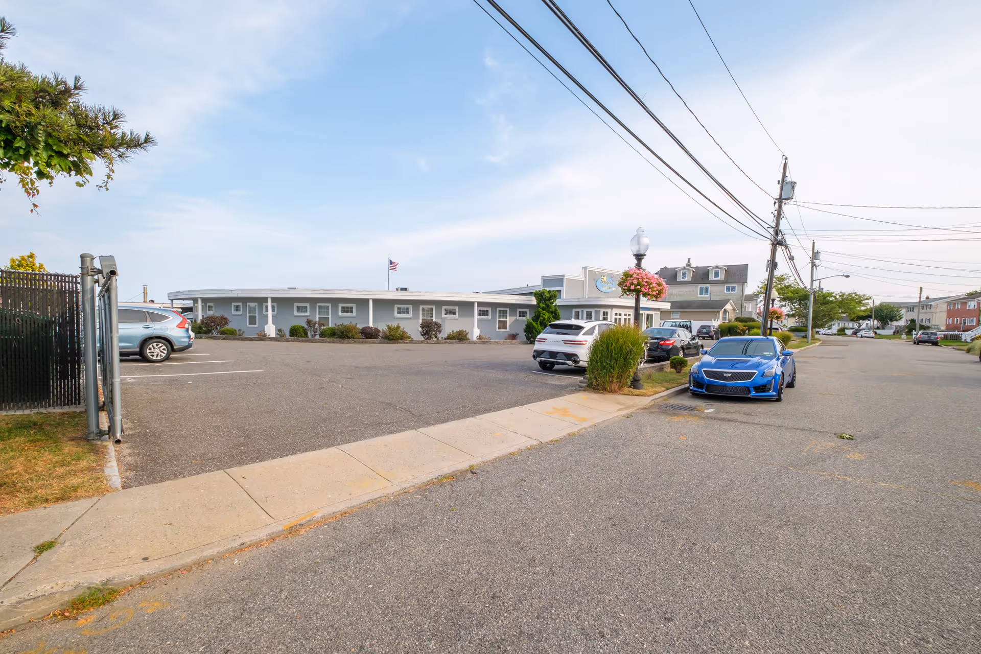 Exterior view of Babylon Beach House facility with a parking lot in front, several parked cars, a sidewalk, and a street with utility poles and wires under a partly cloudy sky.