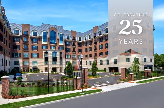 Exterior view of a large multi-story brick and beige assisted living facility building with a gated entrance, landscaped greenery, and a clear blue sky. A translucent overlay on the right side of the image reads 'Celebrating 25 Years'.