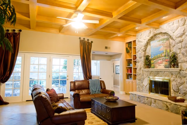 A cozy living room with a wooden coffered ceiling and a ceiling fan. The room features two brown leather sofas arranged around a dark wooden coffee table with drawers. There is a large stone fireplace with a colorful painting above it and built-in wooden shelves on the side. French doors with glass panes lead to an outdoor patio area. The room is warmly lit and has brown curtains on the windows.