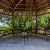 View of a wooden gazebo with lattice sides in an outdoor garden area. Inside the gazebo, there is a round table with four chairs arranged around it. Green trees and grass are visible in the background.