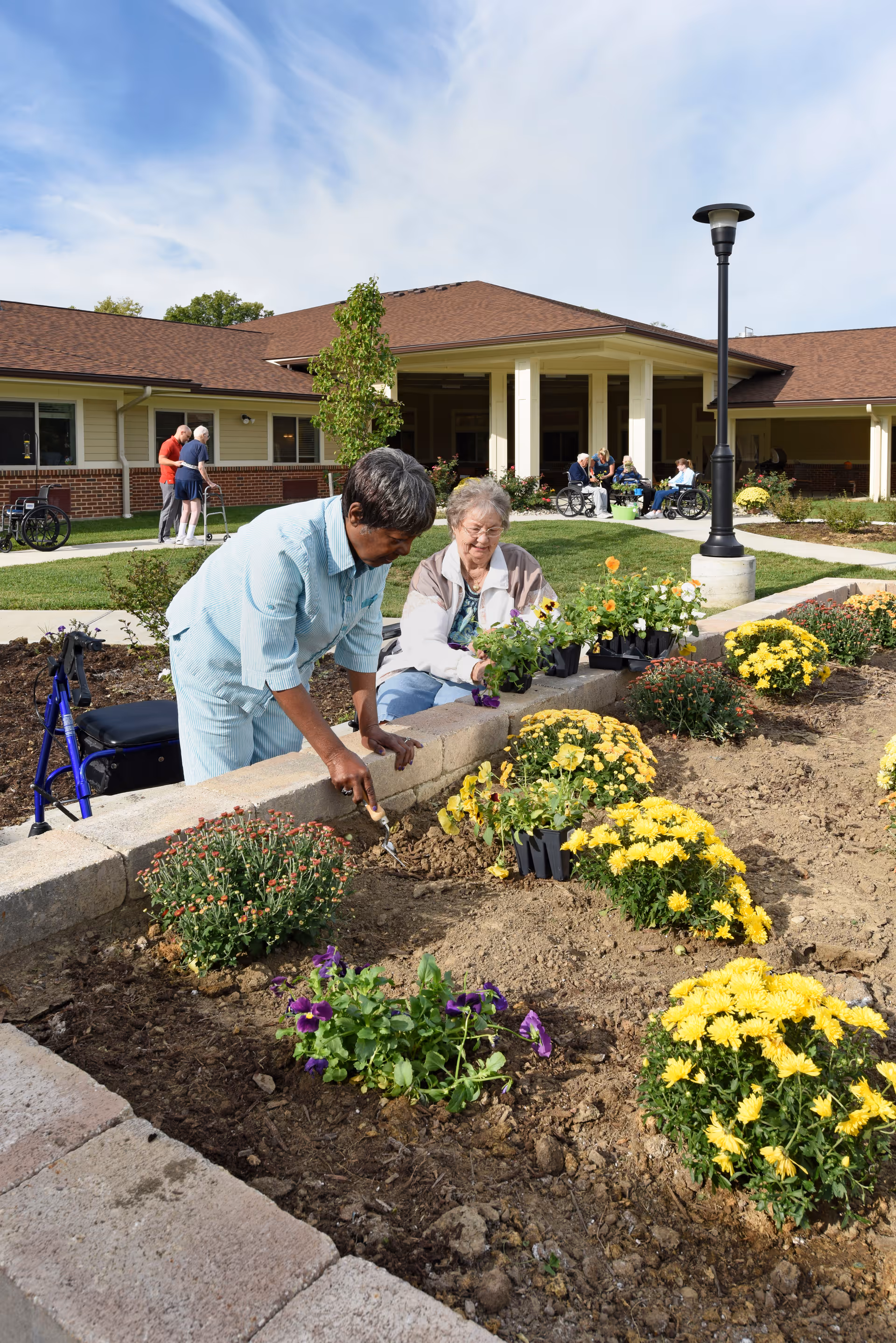 Two elderly women gardening in a flower bed outside a nursing center building. One woman is using a small gardening tool to dig in the soil while the other holds a potted plant. Other elderly people and caregivers are visible in the background near the building entrance and on the lawn.