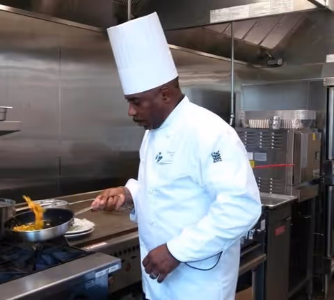 A chef wearing a white chef's coat and tall white chef hat is cooking food in a pan on a stove in a commercial kitchen.